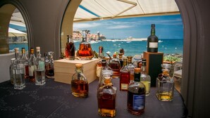 A table with various bottles of liquor, a wooden box, and a bottle of wine, viewed through a window overlooking a coastal town and the sea. - Le Relais des trois Mas (Collioure)