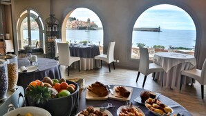 A dining area with tables set for a meal, a fruit bowl, and a view of the sea through arched windows. - Le Relais des trois Mas (Collioure)