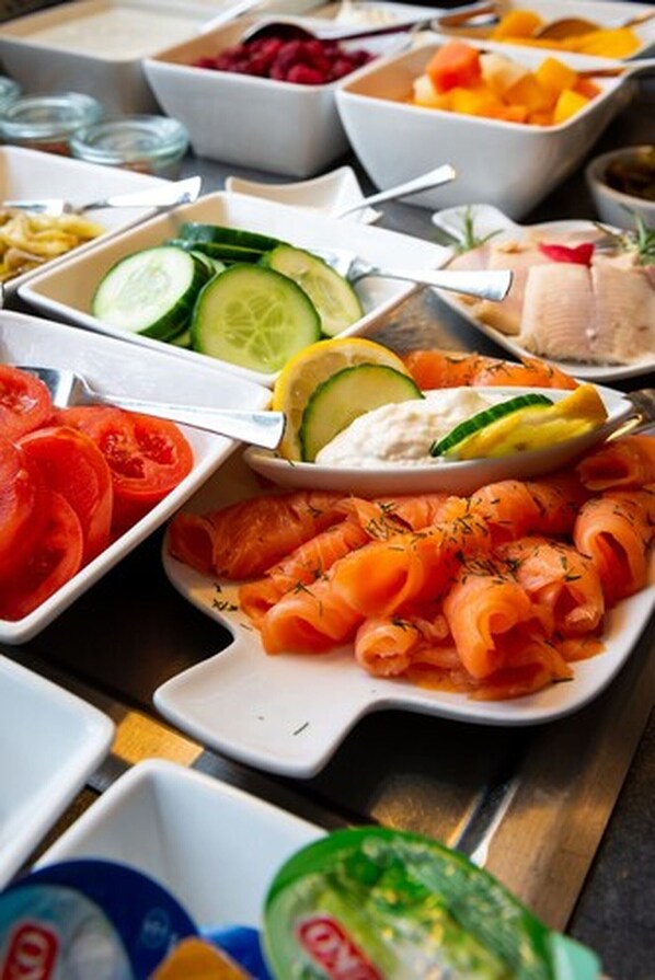 Un bufé con diversos alimentos, incluyendo salmón ahumado, tomates, pepinos y fruta.