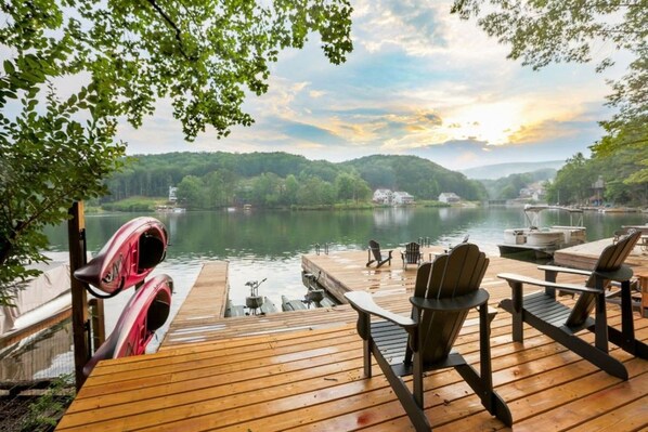 A wooden deck with chairs and a kayak, overlooking a calm lake with boats and houses in the distance.