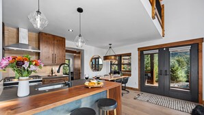 A modern kitchen with wooden cabinets, a central island with stools, pendant lighting, and a view through large sliding doors. - End Unit  - EV Charger, Fireplace & Jacuzzi (Estes Park)