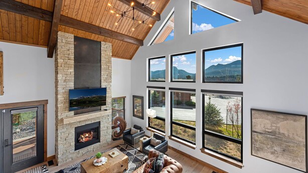 A spacious living room with a stone fireplace, large windows offering mountain views, and a high wooden ceiling. - End Unit  - EV Charger, Fireplace & Jacuzzi (Estes Park)