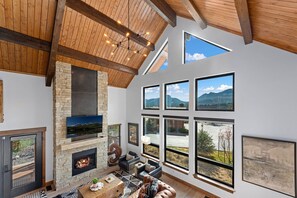 A spacious living room with a stone fireplace, large windows offering mountain views, and a high wooden ceiling. - End Unit - EV Charger, Fireplace & Jacuzzi (Estes Park)