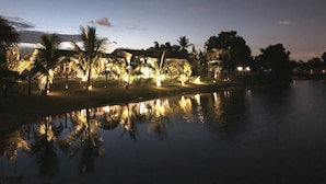 A nighttime view of a waterfront property with a reflective pool, palm trees, and illuminated buildings.