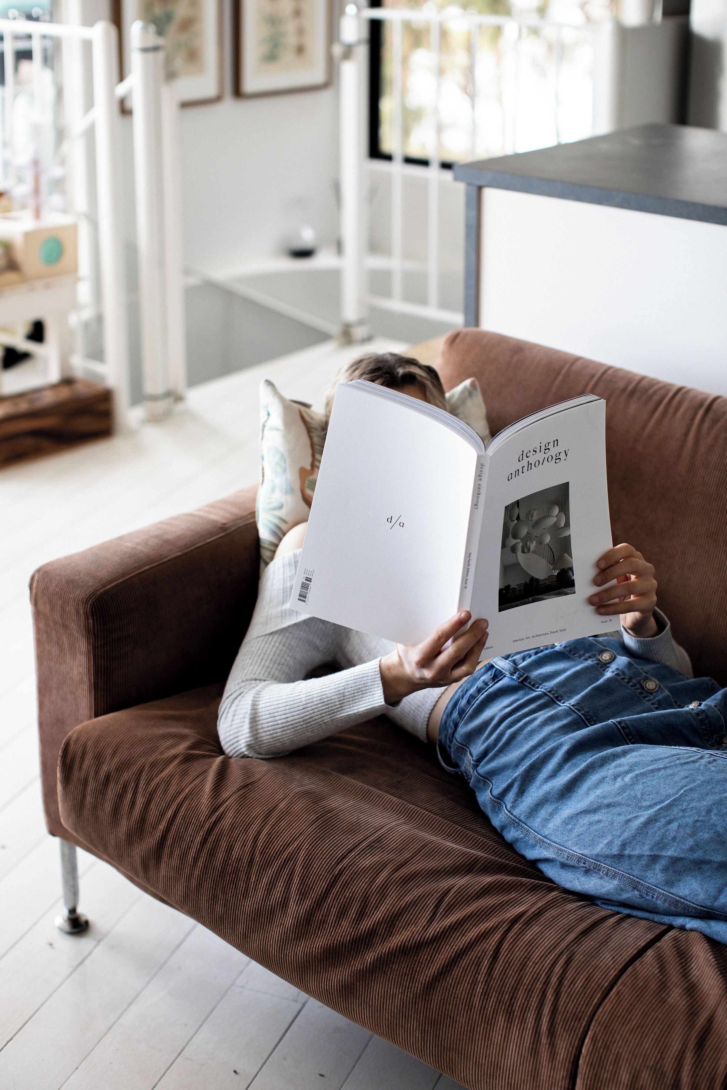 A person reading a book on a brown couch in a well-lit room with a white railing and a view of the outdoors.