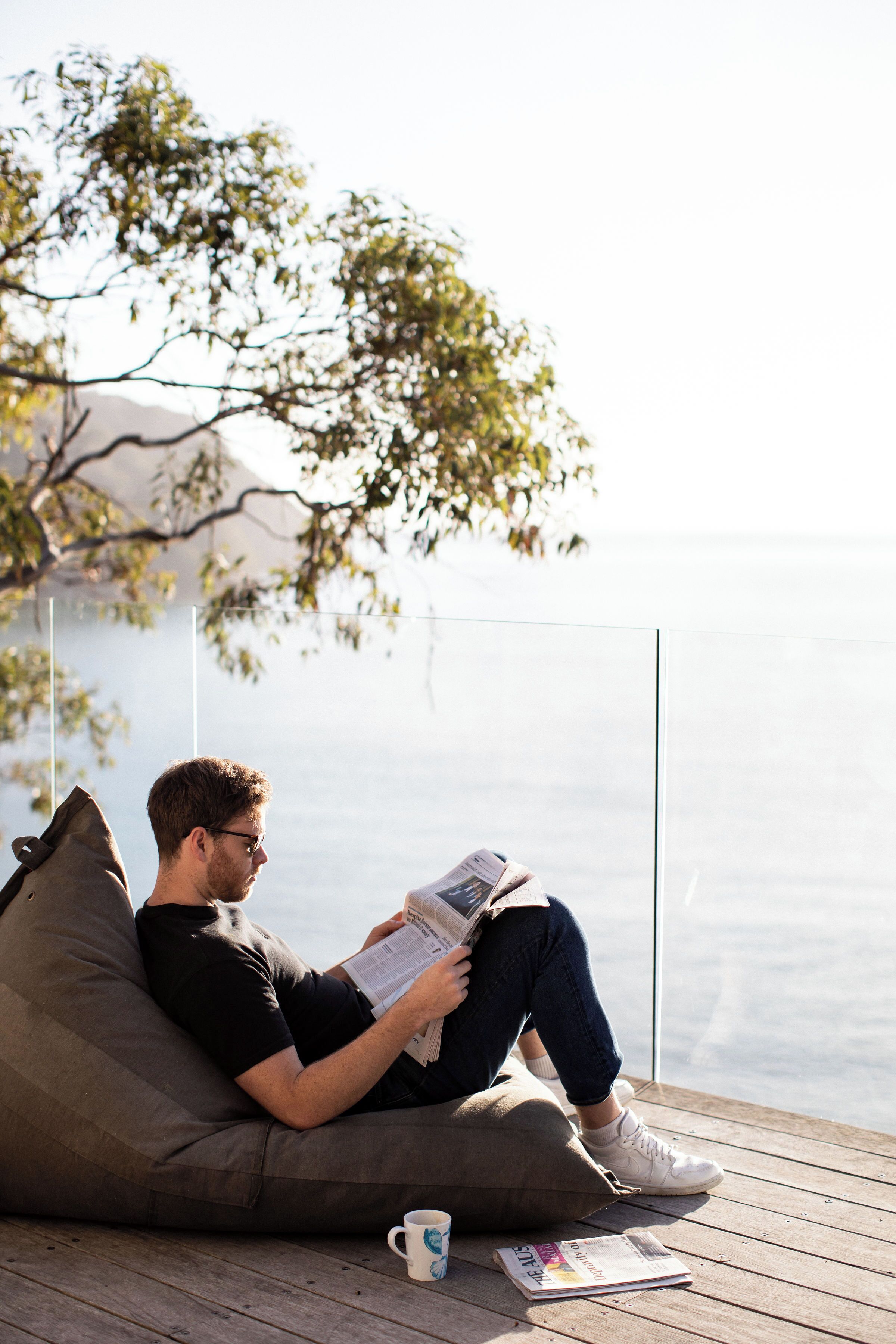 A person sitting on a bean bag chair reading a book on a wooden deck with a clear view of the water in the background.