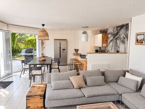 A modern living room with a sectional sofa, a dining area with a table and chairs, a kitchen with a refrigerator and a microwave, and a large window providing a view of the outdoors.