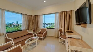 A hotel room with a sofa, armchairs, a coffee table, a TV, and large windows with curtains.