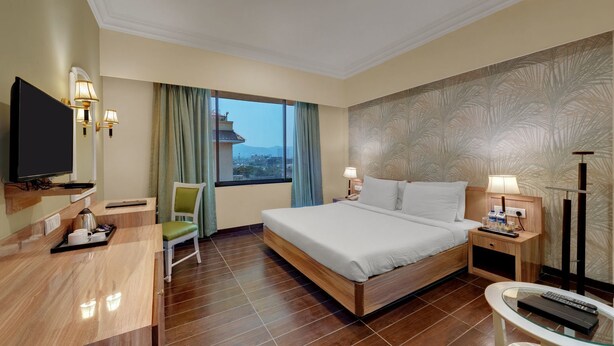 A hotel room with a bed, bedside tables, a TV, a desk with a chair, a window with curtains, and a view of a cityscape.