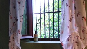 Ein Zimmer mit einem Fenster, das mit floralen Vorhängen drapiert ist, einem kleinen Holzregal und einem runden Hocker mit gemustertem Sitz.
