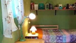 Ein kleines, gemütliches Zimmer mit einem Holztisch, einer Lampe, einem Bett mit gemustertem Überwurf und einem Regal mit diversen Gegenständen.