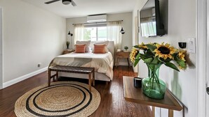 Une chambre à coucher avec un lit, des tables de chevet, un miroir, un ventilateur de plafond, une fenêtre avec des rideaux et un vase rempli de tournesols.