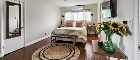 Een slaapkamer met een bed, nachtkastjes, een spiegel, een plafondventilator, een raam met gordijnen en een vaas met zonnebloemen.