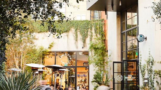 Un espace café en plein air, avec des tables et des parasols, entouré de verdure et bordé d’un bâtiment à la façade en verre.