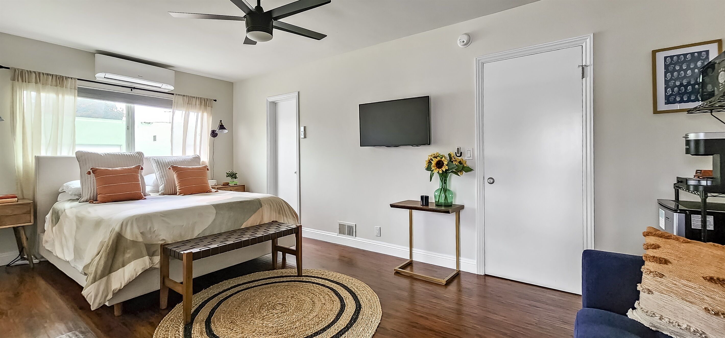 A bedroom with a bed, bedside tables, a TV, a wall-mounted heater, a window with curtains, a small table with a vase, and a chair.