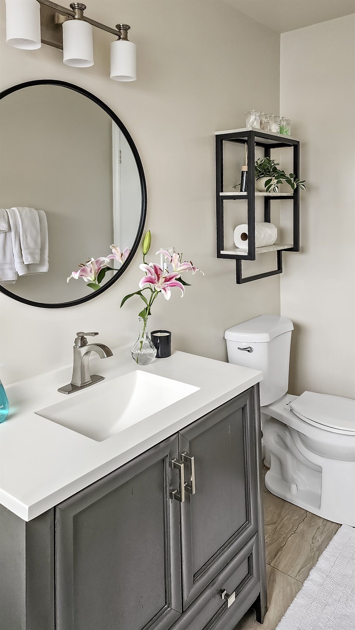 A bathroom with a white sink, dark cabinets, a round mirror, a toilet, and a shelf with toiletries.