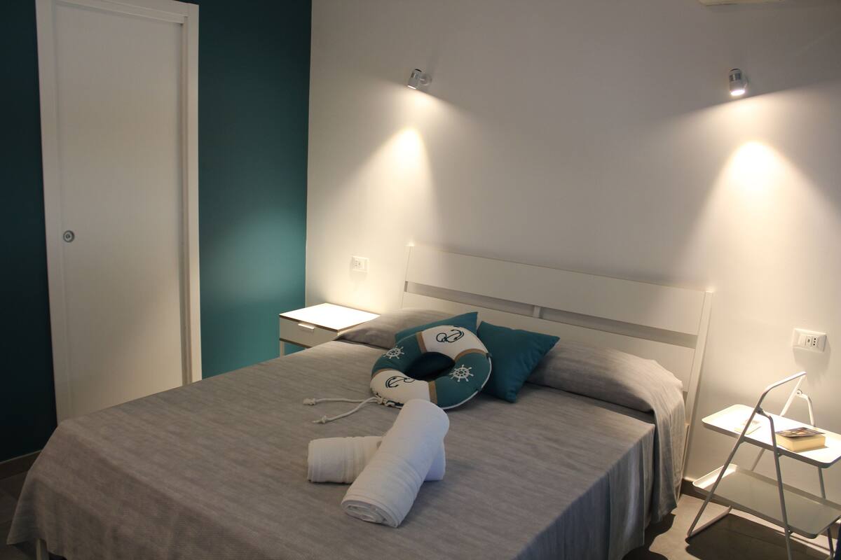A neatly made bed with a teal pillow, a white towel, and a bedside table with a book.