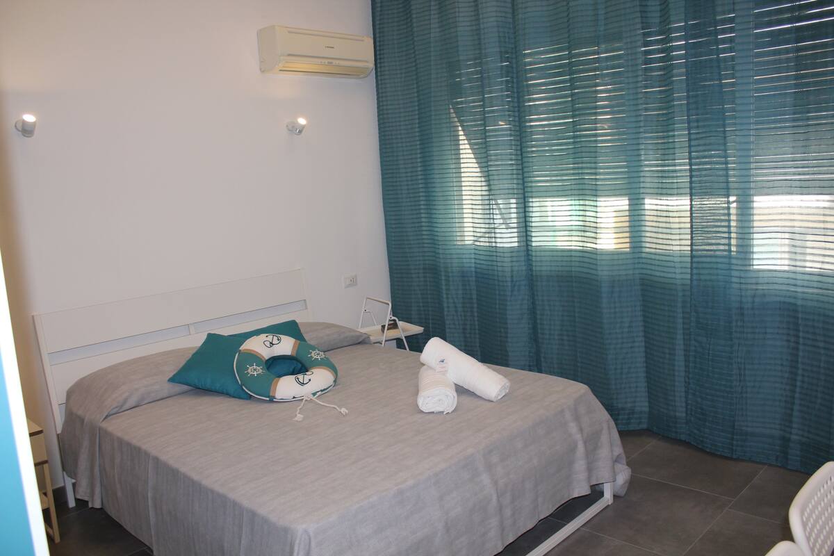 A neatly made bed with a grey bedspread, a teal pillow, and two rolled towels.