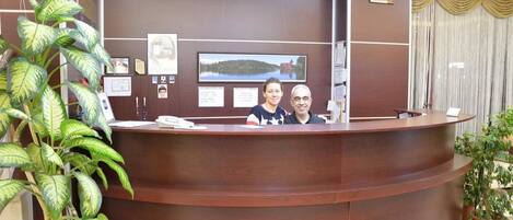 A reception desk with framed pictures and certificates on the wall, a potted plant to the left, and a curtain to the right.