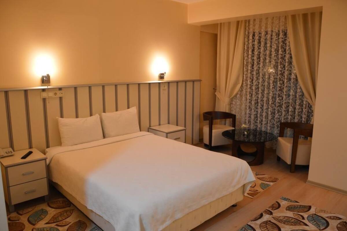 Habitación de hotel ordenada con cama, mesitas de noche, una pequeña mesa con silla y una ventana con cortinas.