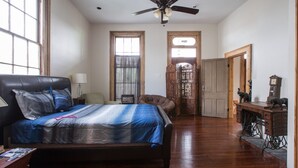Ein Schlafzimmer mit Bett, Schreibtisch, Stuhl, Deckenventilator und einer Tür zu einem weiteren Raum.