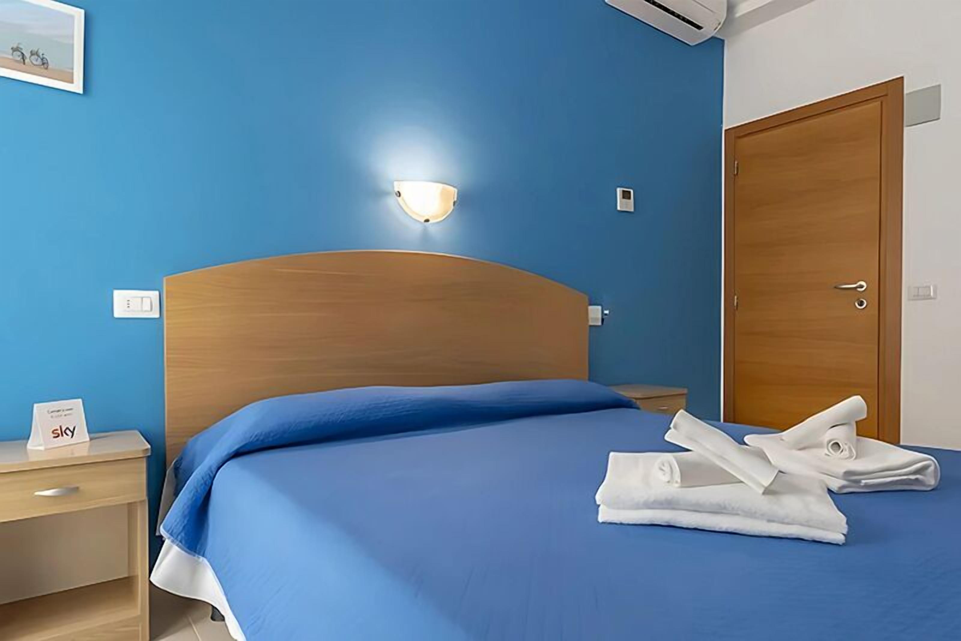 A hotel room with a blue bedspread, a wooden headboard, a nightstand with a 'sky' sign, and a door with a handle.