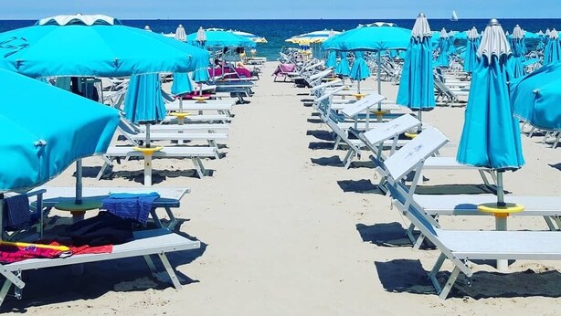 A beach with rows of sun loungers and blue umbrellas, leading to the sea. - Hotel Italy (Misano Adriatico)