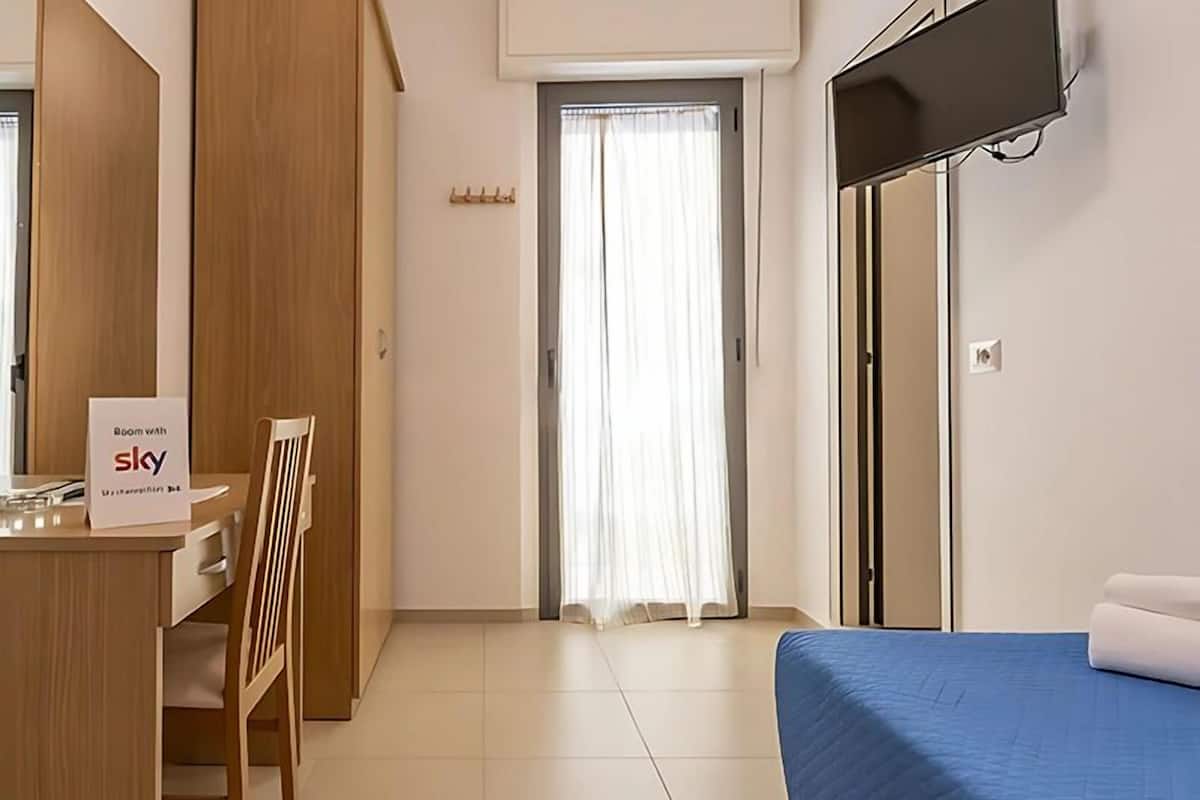 A hotel room with a bed, a desk with a chair, a TV mounted on the wall, and a window with curtains.