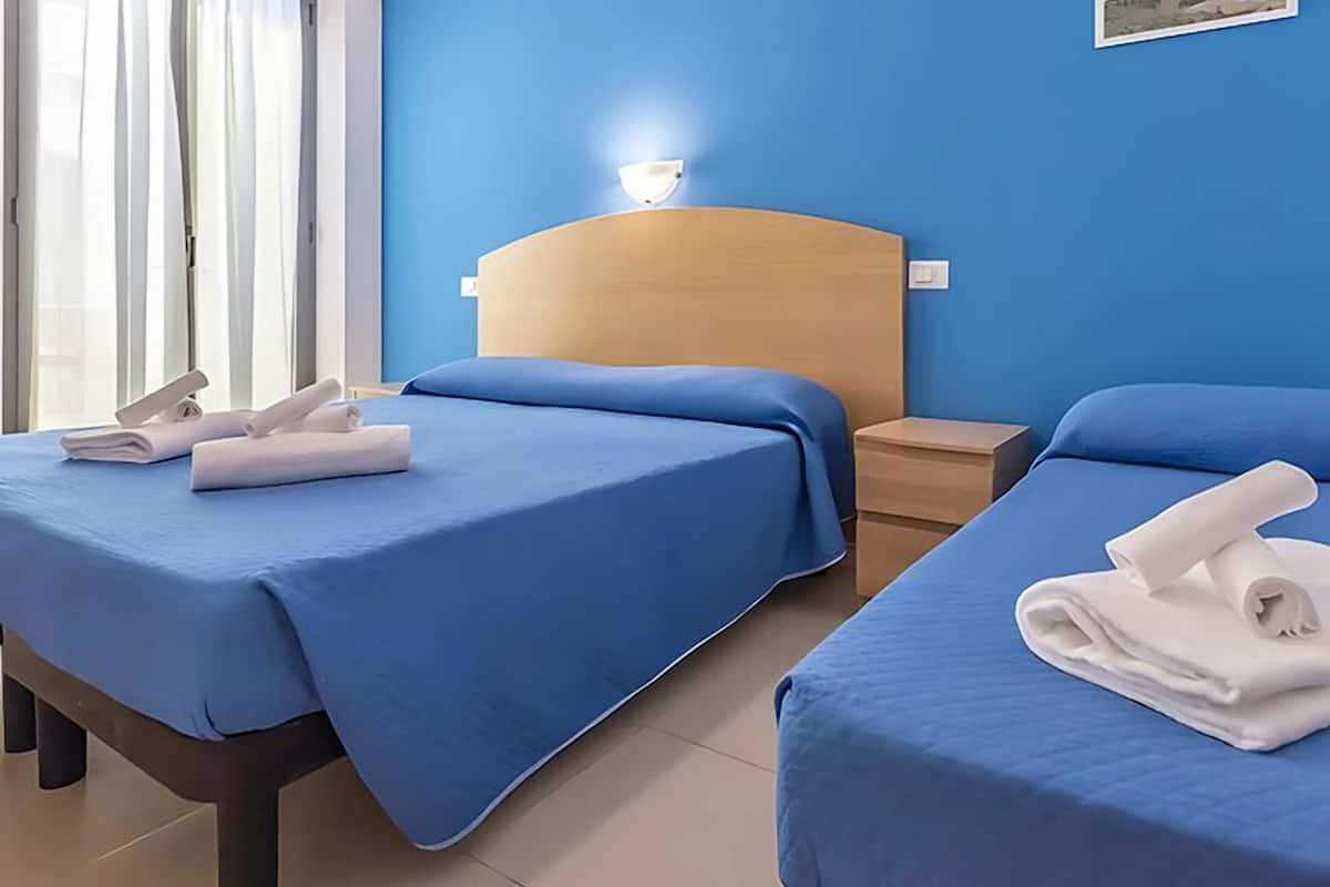 A hotel room with two beds, each with a blue bedspread and white towels. There is a nightstand with a lamp between the beds.