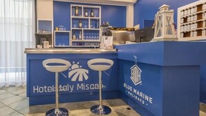 A hotel reception area with a blue counter, two bar stools, a coffee machine, and shelves with bottles and glasses. - Hotel Italy (Misano Adriatico)