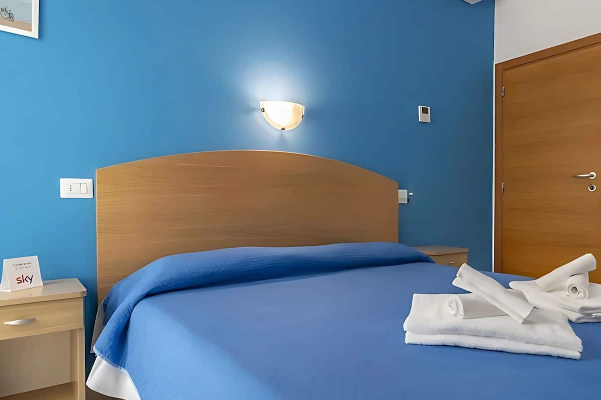 A hotel room with a wooden headboard, a blue bedspread, a nightstand with a lamp, and a door.