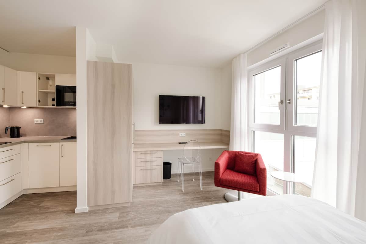 Quarto de hotel moderno com uma cama, escrivaninha, cadeira, televisão e uma janela grande.