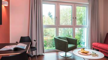 Un salon moderne doté d’une grande fenêtre, d’un fauteuil vert, d’un canapé rouge et d’une petite table ronde sur laquelle sont disposées des bananes et des fleurs.