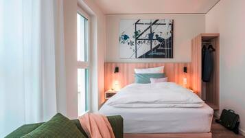 Ein modernes Hotelzimmer mit einem großen Bett, einem grünen Sessel, einem an der Wand befestigten Kunstwerk und einem Holzschrank.