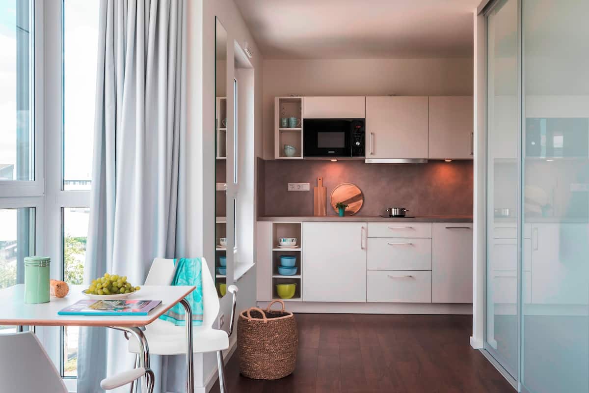 Cozinha compacta com armários brancos, micro-ondas e pia. Há uma área de refeições com mesa e cadeiras, e uma janela com cortinas.