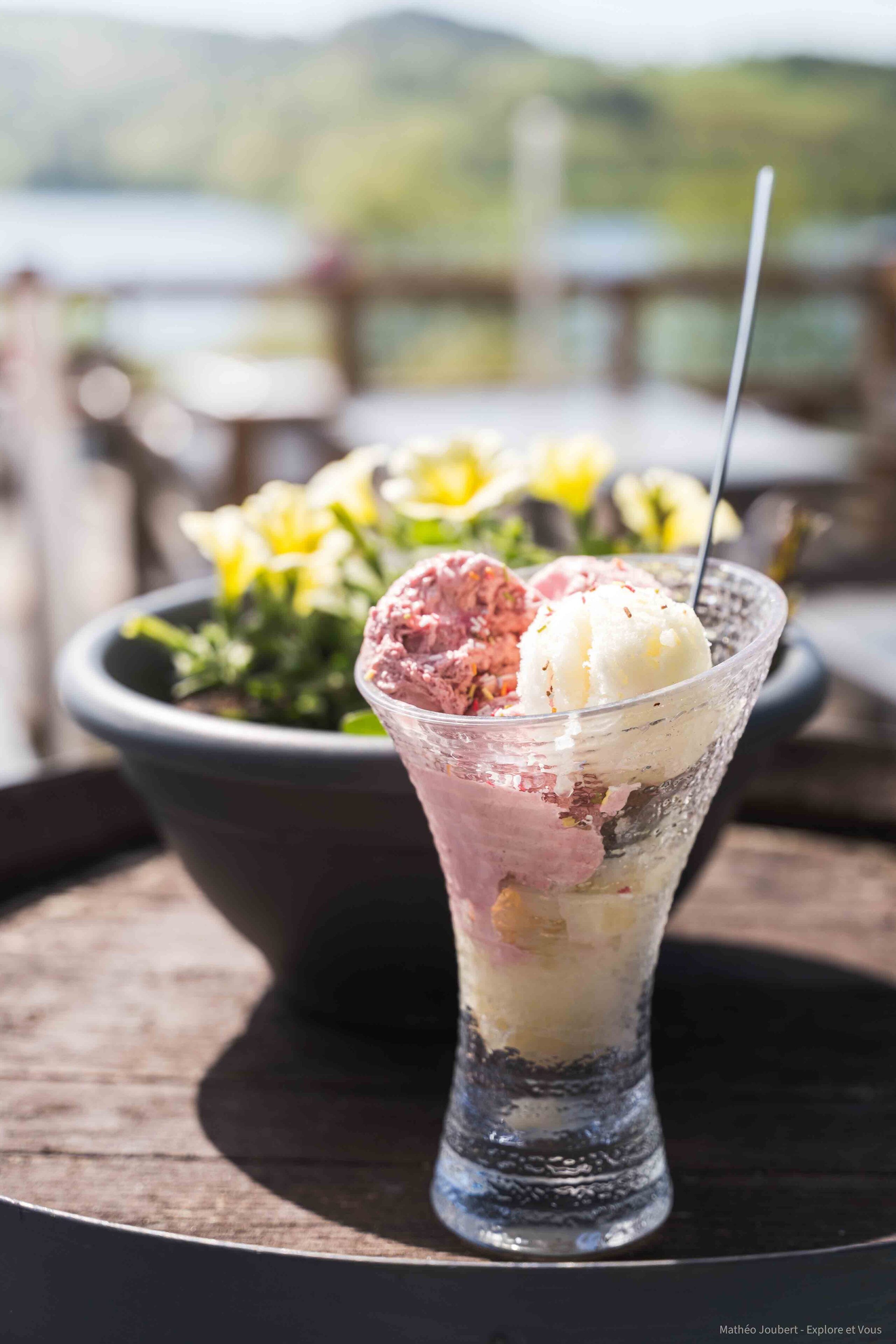Un dessert composé de plusieurs boules de glace servies dans un verre transparent, posé sur une table en bois, avec un arrière-plan extérieur flou.