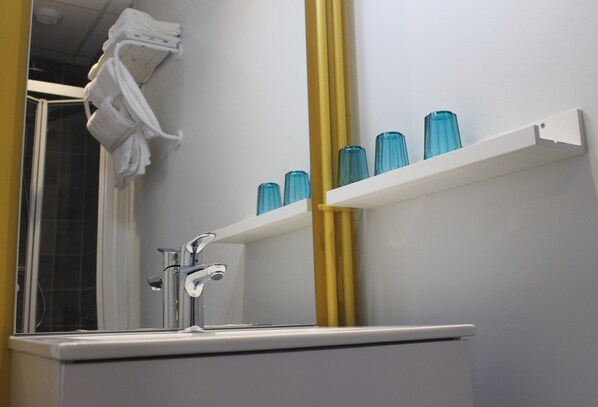 Un baño con lavamanos blanco, grifo cromado y un espejo encima. Hay botellas de vidrio azul en un estante blanco, y un perchero con toallas blancas.