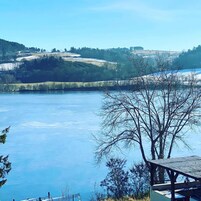 Vista de un lago con muelle de madera y mesa de picnic, rodeado de árboles y colinas.