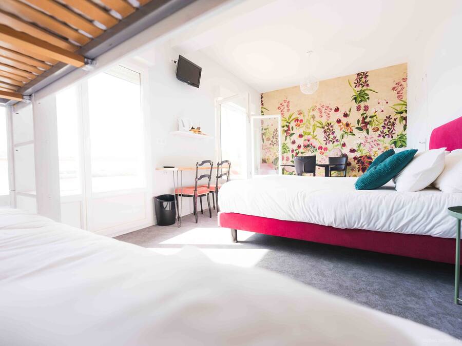 A modern bedroom with a large bed, a desk, and a chair. There is a window providing natural light and a wall with a floral mural.