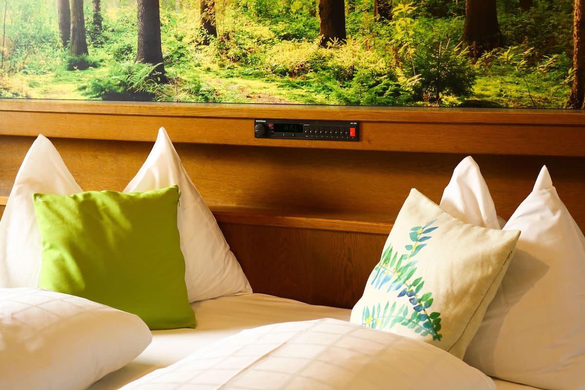 A hotel room with a bed, pillows, and a view of a forest through a window.