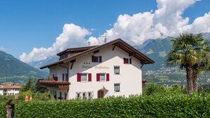Un bâtiment à deux étages doté d’un balcon, entouré d’une végétation luxuriante et avec des montagnes en arrière-plan.
