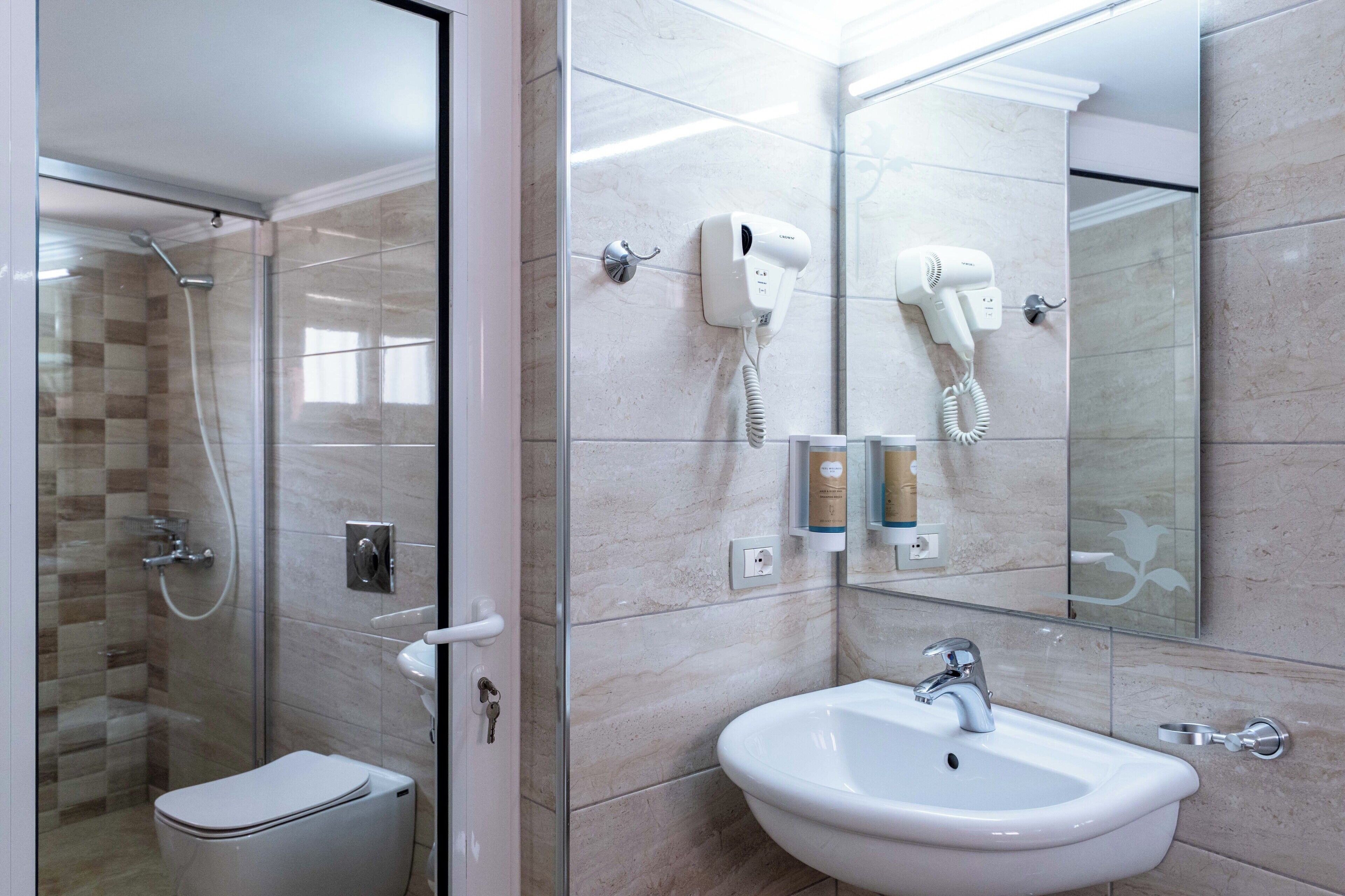 Une salle de bain avec une douche à parois de verre, des toilettes blanches, un lavabo avec un miroir et deux sèche-cheveux fixés au mur.