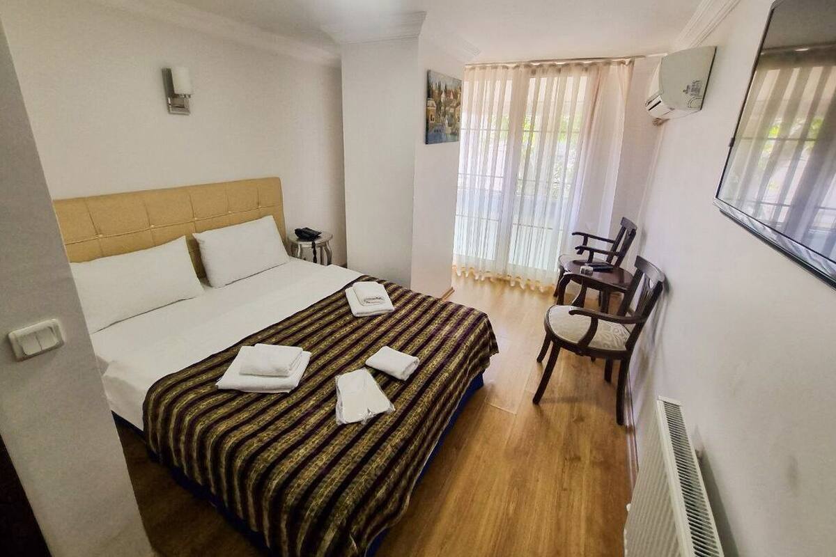 A hotel room with a bed, a desk, and a chair. There is a window with curtains, a radiator, and a wall-mounted air conditioning unit.