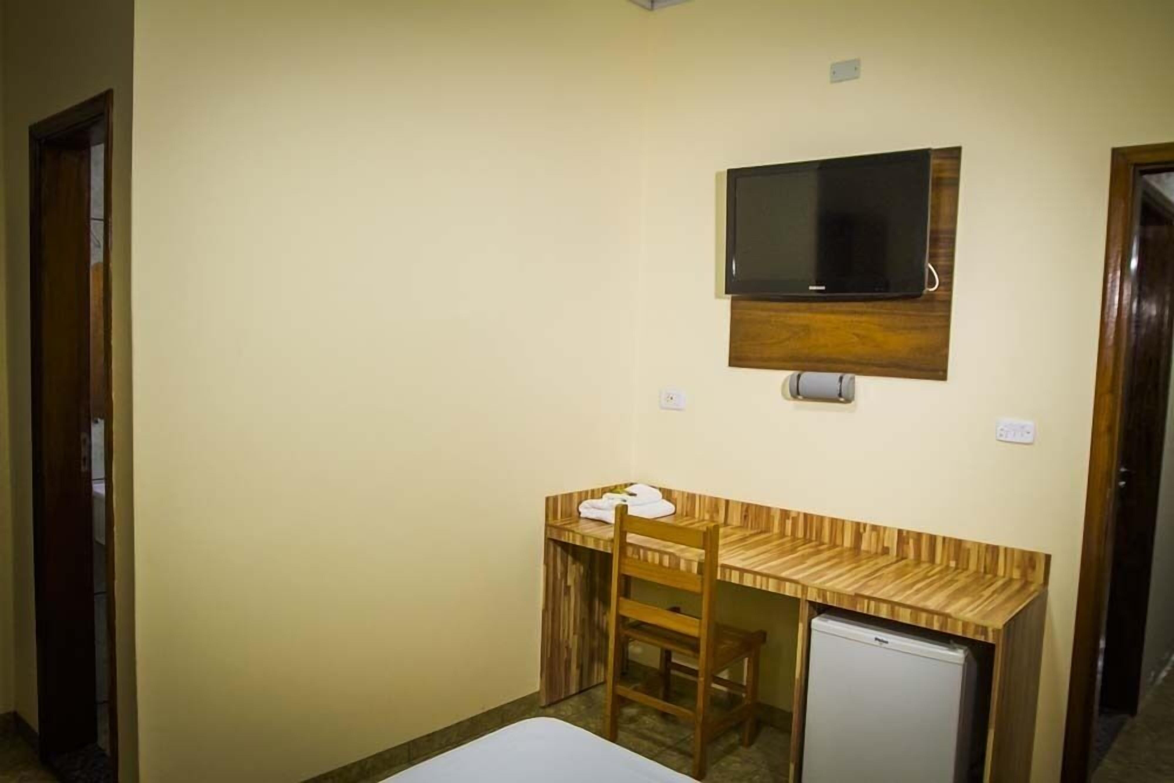 A small room with a wooden desk, a television mounted on the wall, and a small refrigerator.