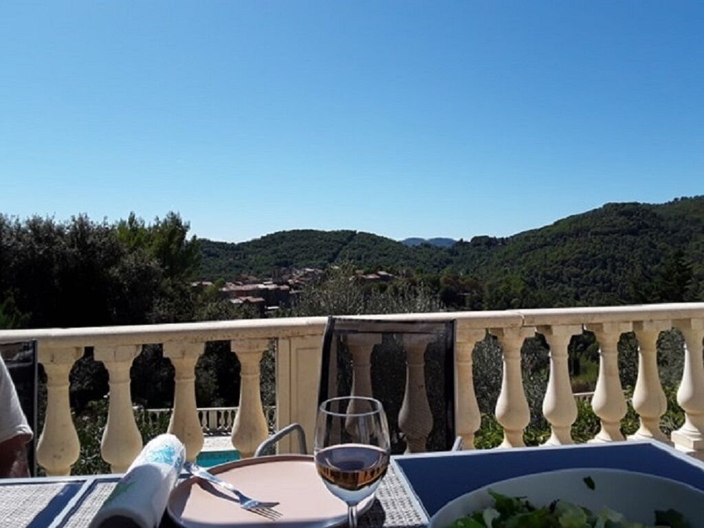 A balcony with a table set for dining, a glass of wine, and a view of a hilly landscape.