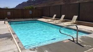 A rectangular swimming pool with clear blue water, surrounded by a wooden fence and a concrete deck. There are lounge chairs along the poolside.