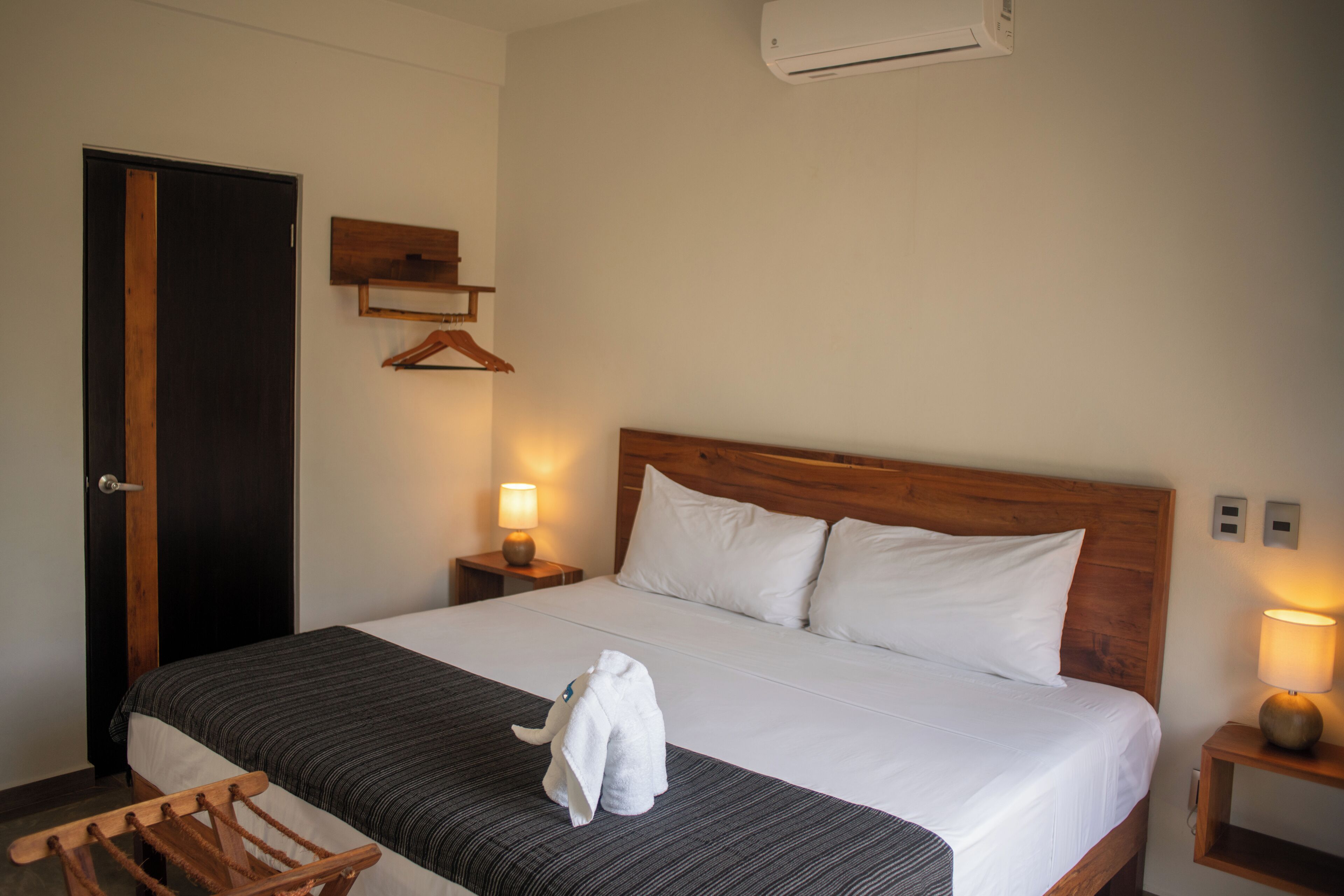 A hotel room with a bed, a wooden headboard, a nightstand with a lamp, a coat rack, and a door.