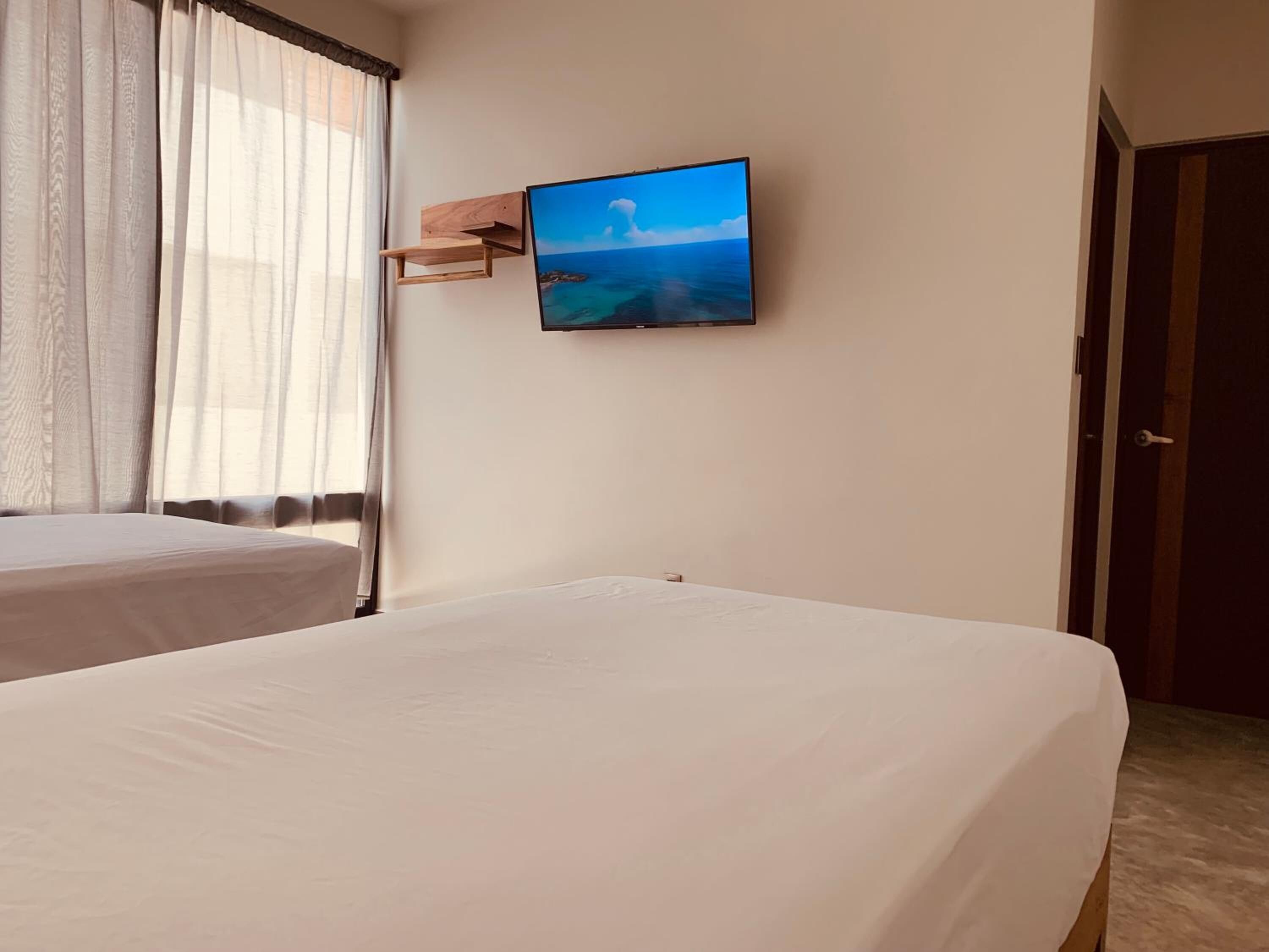 A hotel room with a bed, a TV mounted on the wall, and a window with sheer curtains.
