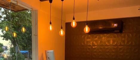 A reception area with a concrete counter, patterned floor tiles, hanging light bulbs, and a large potted plant.