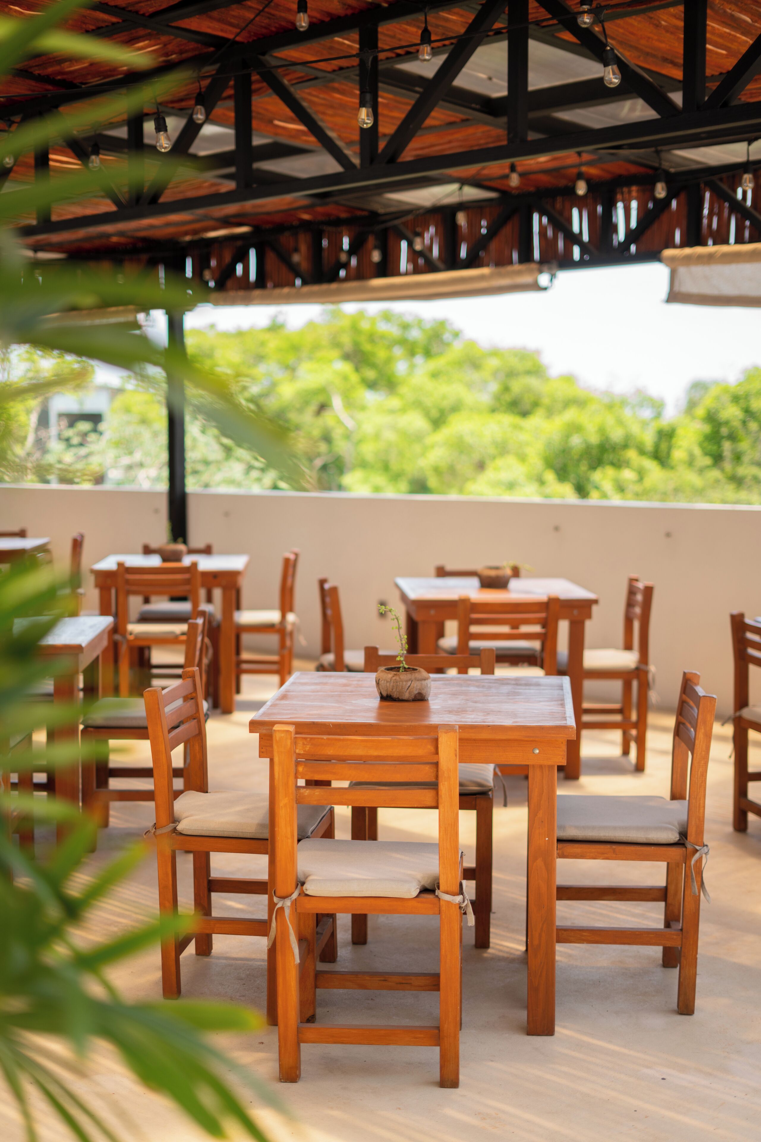 Un patio al aire libre con mesas y sillas de madera, techo de paja y vistas al verdor.